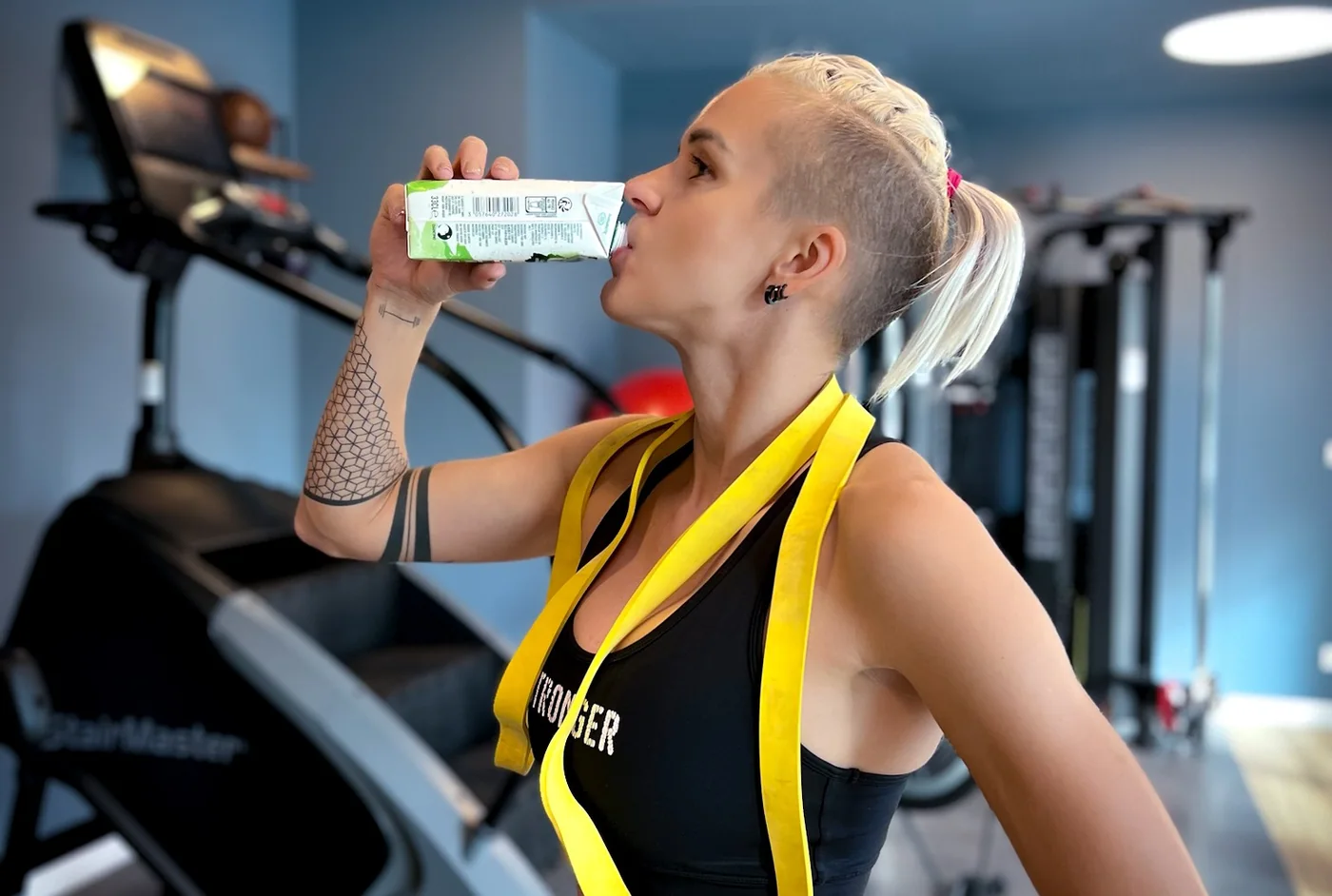 Olga in a gym drinking from a small carton, with a yellow resistance band around her neck