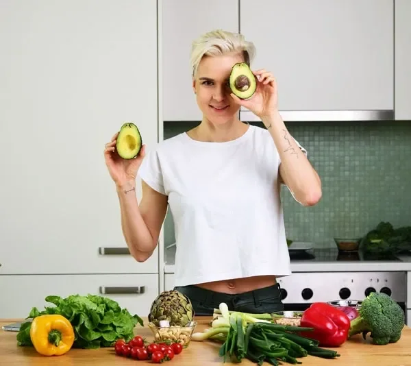 Olga in a kitchen holding up two halves of an avocado, fresh vegetables on the counter